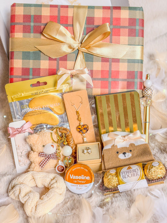 Box cadeau cocooning ourson BloomLov avec accessoires, soins, papeterie et chocolats devant un paquet à carreaux


