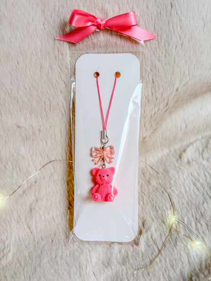 Phone charm BloomLov avec ourson rose et nœud transparent sur une attache rose, présenté sur carte blanche


