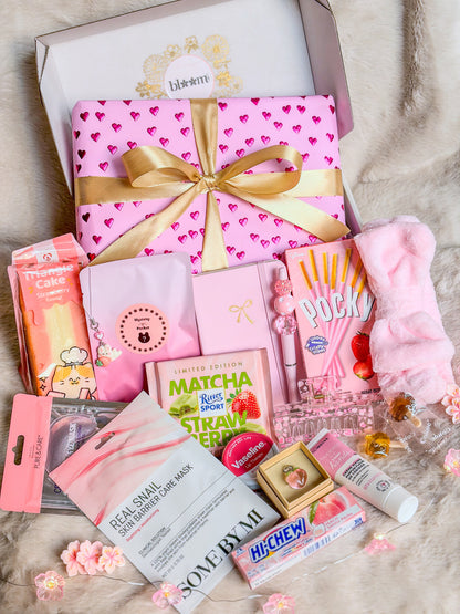 Pinky Self-Care Box BloomLov avec soins, accessoires roses et gourmandises devant un paquet à cœurs et ruban doré